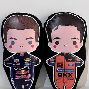 Könnte beinhalten: Zwei schwarze Plüschkissen mit Cartoon-Illustrationen von Formel-1-Fahrern. Ein Kissen zeigt einen Fahrer in einem marineblauen Rennanzug mit Red Bull- und Oracle-Logos. Das andere Kissen hat einen Fahrer in einem orangefarbenen Rennanzug mit Android- und Dell Technologies-Logos.