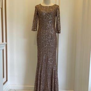 May include: A floor-length, bronze-colored gown with a fitted bodice and a flared skirt. The dress is covered in sequins and has three-quarter sleeves. The dress is displayed on a mannequin in front of a neutral background.