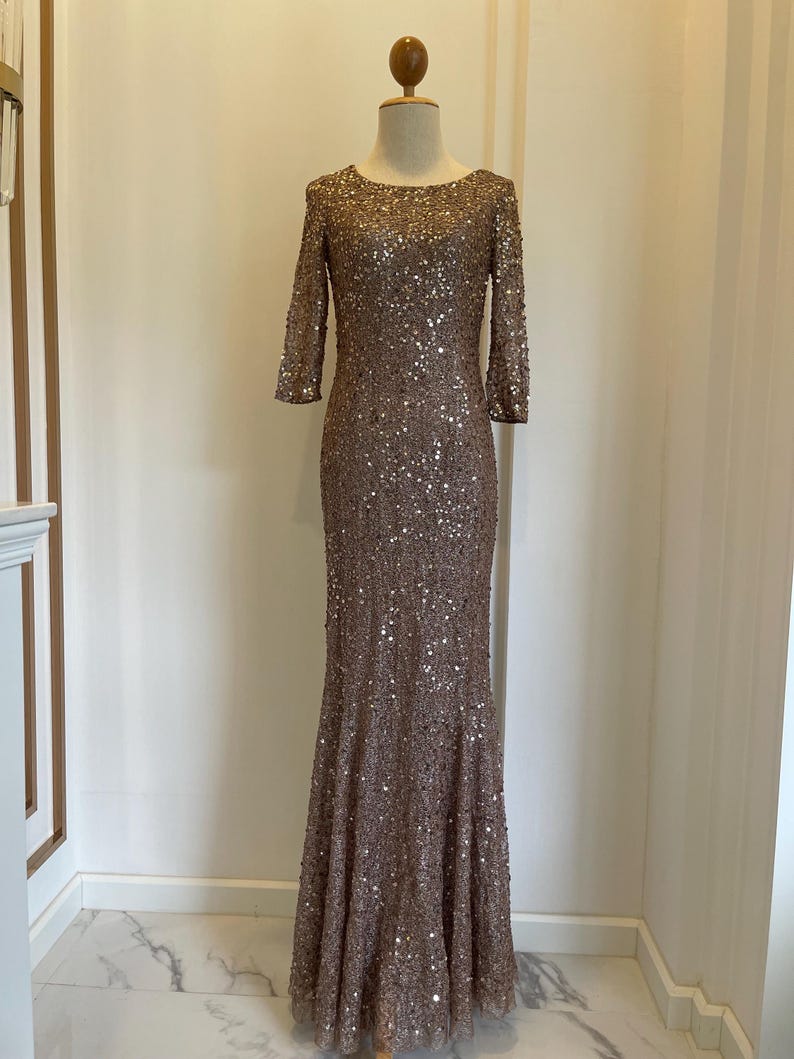 May include: A floor-length, bronze-colored gown with a fitted bodice and a flared skirt. The dress is covered in sequins and has three-quarter sleeves. The dress is displayed on a mannequin in front of a neutral background.