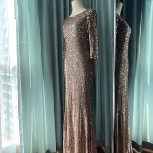 May include: A floor-length, brown sequined gown with long sleeves and a scoop neckline. The dress is displayed on a mannequin in front of a mirror, reflecting the dress's design. The dress has a fitted bodice and a flared skirt.