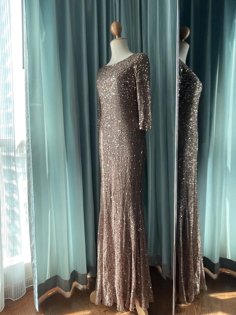 May include: A floor-length, brown sequined gown with long sleeves and a scoop neckline. The dress is displayed on a mannequin in front of a mirror, reflecting the dress's design. The dress has a fitted bodice and a flared skirt.
