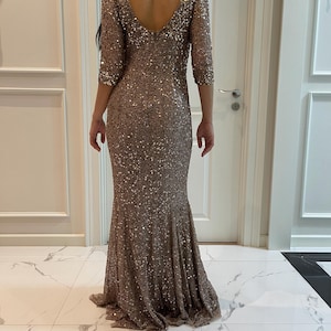 May include: A floor-length, brown sequined gown with a V-neckline and three-quarter sleeves. The dress has a fitted bodice that flares out at the hem. The back of the dress has a low V-cut. The dress is covered in small, shimmering sequins.