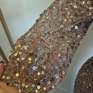 May include: Close-up of a sleeve covered in bronze-colored sequins. The sequins are round and reflect light, creating a shimmering effect. The fabric underneath appears to be a fine mesh, adding texture and depth to the design. The sleeve is part of a garment.