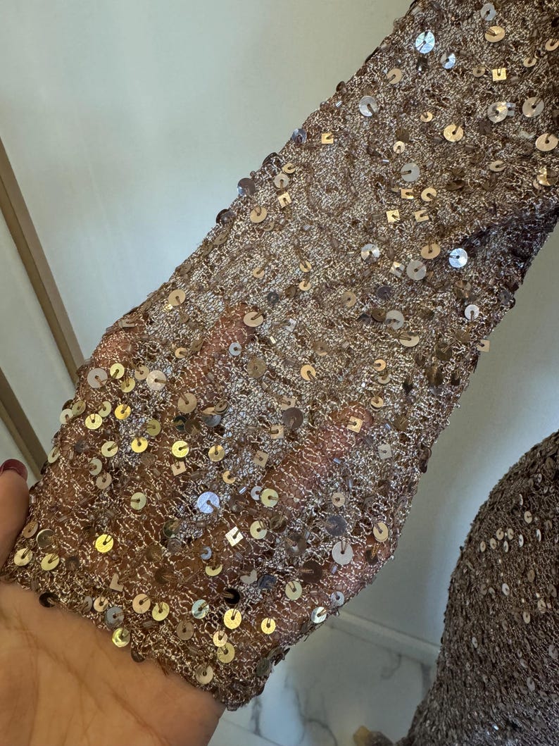 May include: Close-up of a sleeve covered in bronze-colored sequins. The sequins are round and reflect light, creating a shimmering effect. The fabric underneath appears to be a fine mesh, adding texture and depth to the design. The sleeve is part of a garment.