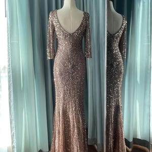 May include: A floor-length, bronze-colored sequined gown with a low back and three-quarter sleeves. The dress is displayed on a mannequin, with a matching reflection in a nearby mirror. The dress has a mermaid silhouette.