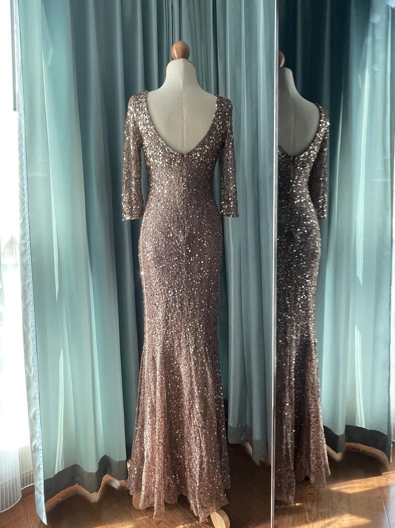 May include: A floor-length, bronze-colored sequined gown with a low back and three-quarter sleeves. The dress is displayed on a mannequin, with a matching reflection in a nearby mirror. The dress has a mermaid silhouette.