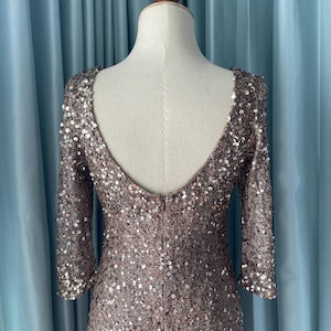 May include: A bronze-colored, sequined dress with a low scoop back and three-quarter sleeves. The dress is displayed on a mannequin, showcasing its form-fitting design. The sequins provide a shimmering effect, ideal for formal events.