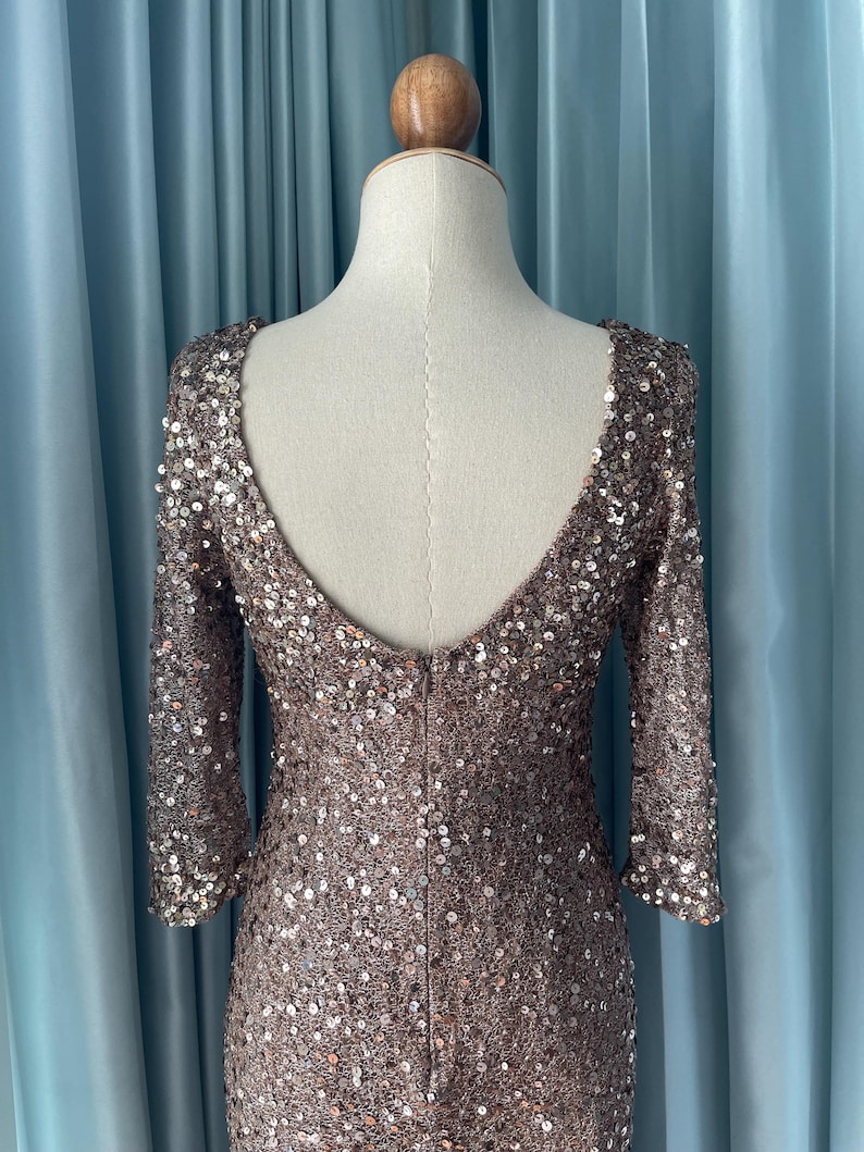 May include: A bronze-colored, sequined dress with a low scoop back and three-quarter sleeves. The dress is displayed on a mannequin, showcasing its form-fitting design. The sequins provide a shimmering effect, ideal for formal events.