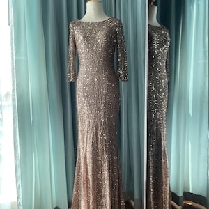 May include: A floor-length, bronze-colored sequined gown with three-quarter sleeves is displayed on a mannequin. The dress has a boat neckline and a fitted silhouette that flares out slightly at the bottom. The dress is reflected in a nearby mirror.