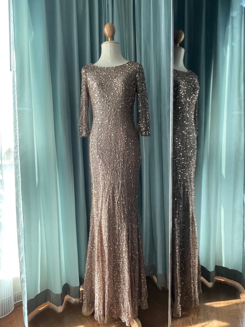 May include: A floor-length, bronze-colored sequined gown with three-quarter sleeves is displayed on a mannequin. The dress has a boat neckline and a fitted silhouette that flares out slightly at the bottom. The dress is reflected in a nearby mirror.