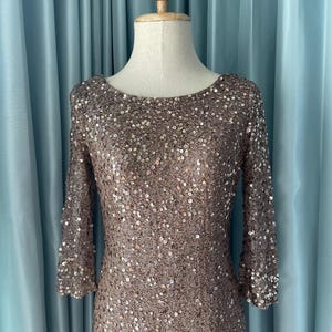 May include: A bronze-colored, sequined dress with three-quarter sleeves. The dress is displayed on a mannequin against a light blue curtain backdrop. The dress has a scoop neckline and is covered in small, reflective sequins.