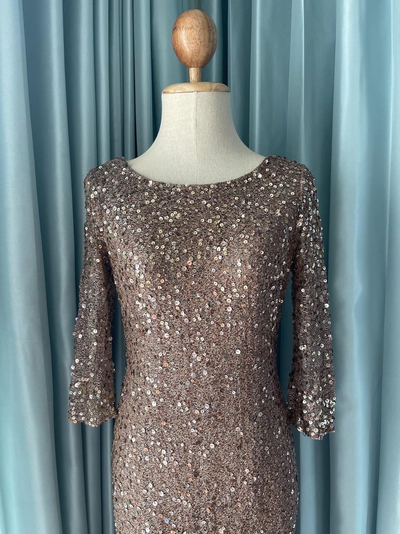 May include: A bronze-colored, sequined dress with three-quarter sleeves. The dress is displayed on a mannequin against a light blue curtain backdrop. The dress has a scoop neckline and is covered in small, reflective sequins.