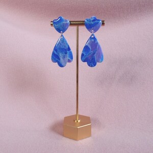 May include: A pair of blue marbled earrings with a floral design. The earrings are displayed on a gold-coloured earring stand with a hexagonal base. The earrings have a heart-shaped top and a teardrop-shaped bottom.