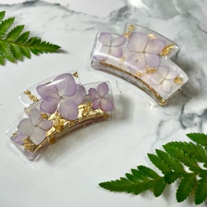 May include: Two clear hair clips with a gold metal bar and pressed purple flowers. The flowers are delicate and have a light, airy feel.