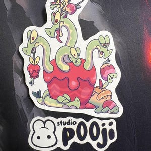 Peut inclure: Autocollant avec une illustration de dessin animé d'un dragon vert à plusieurs têtes émergeant d'une pomme rouge. L'autocollant comprend également le texte "studio pooji" en lettres noires.