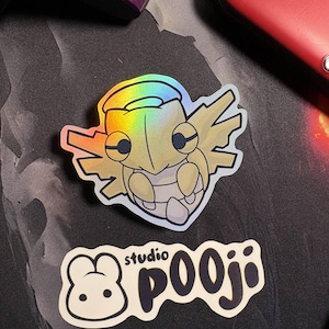 May include: Holographic sticker featuring a stylized insectoid creature with a light yellow body and iridescent highlights. A second sticker with the text "studio pooji" and a cartoon bunny is also visible.