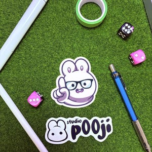 May include: An assortment of stationery and accessories on a green surface. Includes a white pen, a roll of green washi tape, two dice, a blue mechanical pencil, and two stickers with a cartoon bunny design and the text "studio pooji".