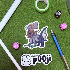 May include: A sticker featuring a fantasy creature with a brown head, gray and purple body, and blue accents. The image also includes a white pen, a blue mechanical pencil, two pink dice, a black die, and a roll of green tape. The sticker has the text "studio POOJI".