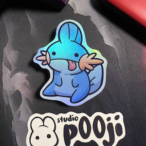 May include: A holographic sticker featuring a cartoon Mudkip character with blue and light brown accents. The sticker has a shiny, iridescent finish. Below the Mudkip sticker is a white sticker with the text "studio pooji" in a black, bold font.