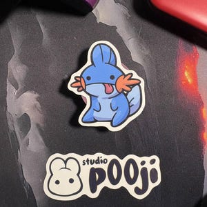 May include: Two stickers on a dark surface. One sticker features a blue cartoon character with orange accents and a red tongue. The second sticker has a white background with a cartoon bunny and the text "studio pooji" in black.