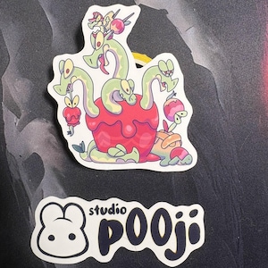 May include: Two stickers on a dark surface. One sticker features a cartoon illustration of a multi-headed green serpent emerging from a red fruit. The other sticker has a white bunny face and the text "studio pooji".