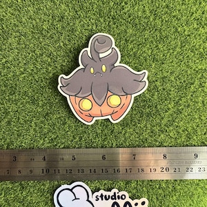May include: A sticker featuring a cartoon character with a pumpkin-like body and a dark gray head. The character has yellow eyes and a smiling mouth. A ruler is placed next to the sticker. A second sticker with a white bunny and the text "studio pooji" is also visible.