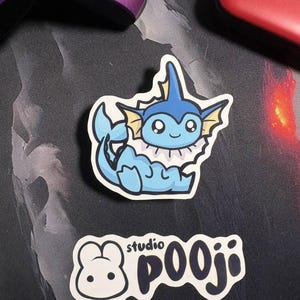 May include: A die-cut sticker featuring a cartoon Vaporeon character in shades of blue, with a white frill and yellow accents. Below is a sticker with a cartoon bear and the text "studio POOJI" in a playful font. The stickers are on a dark surface.