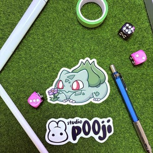 May include: A collection of stationery and accessories on a green surface. Includes a Bulbasaur sticker, a Studio Pooji sticker, a roll of green tape, two dice, a blue mechanical pencil, and two white styluses.
