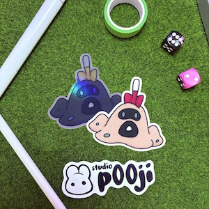 May include: Two stickers with a cartoon character design, one in dark blue and the other in tan, with a red bow. The stickers are on a green surface with a white pen, green tape, and dice. The text "studio pooji" is also visible.