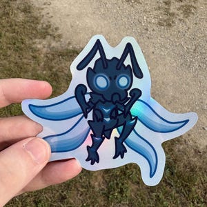 May include: A holographic sticker featuring a stylized, dark blue insect-like creature with large, glowing blue eyes and wings. The sticker has a unique shape and is held against a blurred outdoor background.