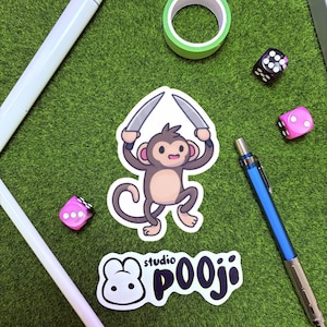 May include: An assortment of stationery and accessories on a green surface. Includes a monkey sticker holding knives, a sticker with the text "studio POOJI", a roll of green tape, two dice, a blue mechanical pencil, and two white styluses.