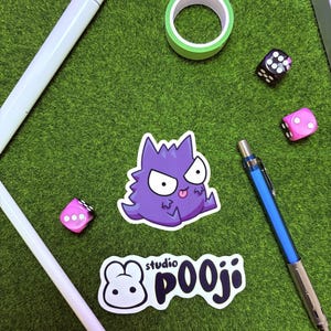 May include: A collection of stationery and accessories on a green surface. Includes a purple Gengar sticker, a white and black "studio POOJI" sticker, a green washi tape roll, two dice, and two pens.
