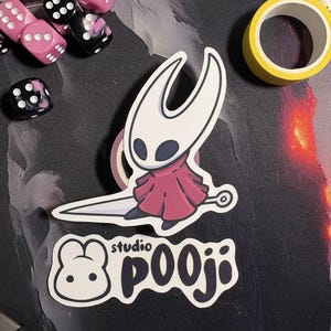 May include: A white and red sticker featuring a character from the video game Hollow Knight, with a sword. Below is a sticker with a bunny logo and the text "studio pooji". Pink and black dice and yellow tape are also visible.
