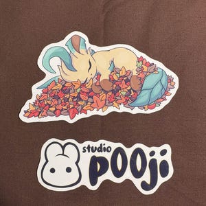 May include: A sticker featuring a cartoon creature sleeping in a pile of autumn leaves. The creature is tan with green and brown accents. Below is a second sticker with the text "studio pooji" and a cartoon bunny.