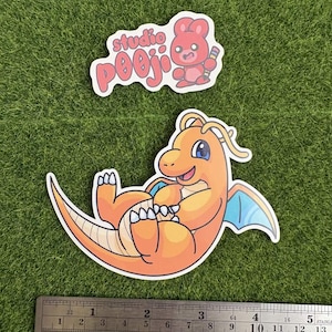 May include: Two stickers on a green surface. One sticker features a red bear holding a pencil with the text "studio pooji". The other sticker is an orange cartoon dragon with blue wings. A ruler is at the bottom.