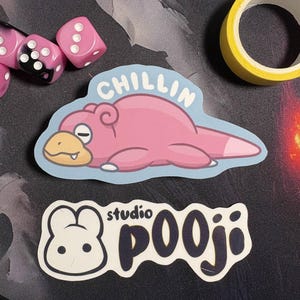 May include: A pink Slowpoke sticker with the word "CHILLIN" above it. The sticker is on a blue background. Below is a sticker with a white bunny head and the words "studio POOJI" in black. Pink dice and yellow tape are also visible.
