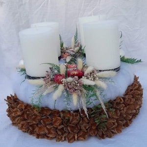 Könnte beinhalten: Ein weißer Adventskranz mit vier weißen Kerzen, verziert mit getrockneten Blumen, Gräsern und Tannenzweigen. Der Kranz steht auf einem Sockel aus braunen, getrockneten Blütenknospen.