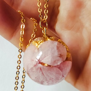 Peut inclure: Un collier à chaîne dorée avec un pendentif sphérique. Le pendentif transparent renferme des pétales de fleurs roses et des paillettes dorées. Le collier est tenu dans une main sur fond blanc.