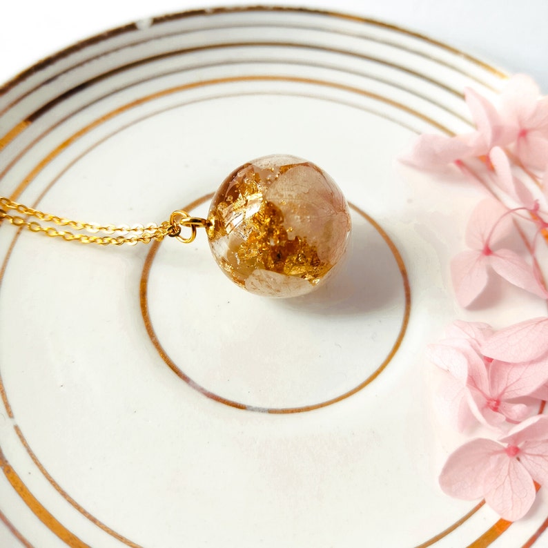 May include: A gold-colored necklace featuring a clear, spherical pendant with gold leaf accents and a delicate chain. The pendant rests on a white plate with gold trim, with pink flowers in the background.