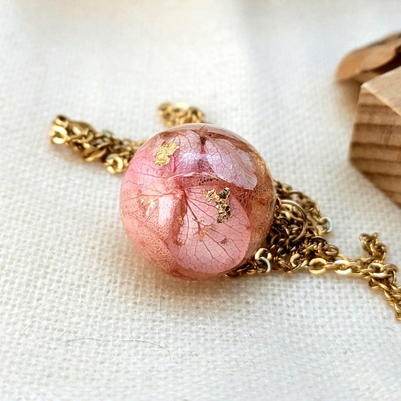 May include: A gold-colored chain necklace with a pendant. The pendant is a clear sphere encasing pink dried flowers and gold leaf flakes. The necklace is displayed on a white surface.