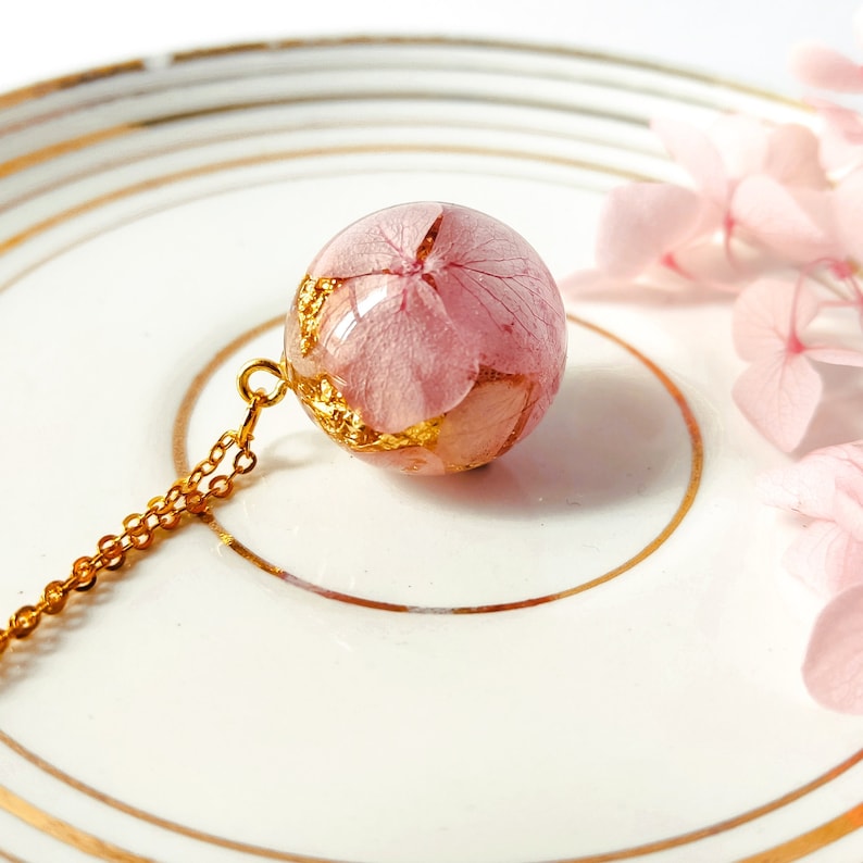 May include: A delicate gold chain necklace featuring a clear, spherical pendant. The pendant encases a pink flower with gold leaf accents. The necklace is displayed on a white plate with a gold rim, with pink flowers in the background.