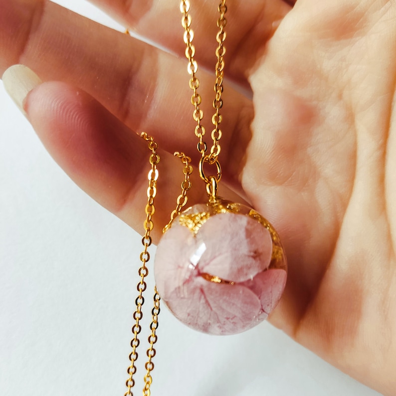 Puede incluir: Un collar de cadena dorada con un colgante redondo que presenta flores prensadas rosas encapsuladas en resina transparente.