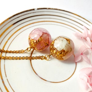 May include: Two round pendant necklaces with gold chains. The pendants are clear spheres filled with pink and white dried flowers and gold leaf flakes. The necklaces rest on a white plate with gold trim.