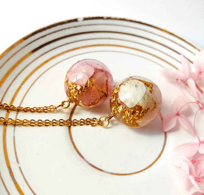 May include: Two round pendant necklaces with gold chains. The pendants are clear spheres filled with pink and white dried flowers and gold leaf flakes. The necklaces rest on a white plate with gold trim.