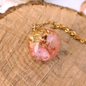 May include: A gold-colored necklace featuring a round pendant. The pendant is a clear sphere filled with pink dried flowers and gold leaf flakes. The necklace rests on a wooden surface.