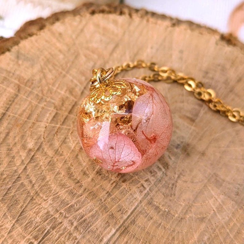 May include: A gold-colored necklace featuring a round pendant. The pendant is a clear sphere filled with pink dried flowers and gold leaf flakes. The necklace rests on a wooden surface.