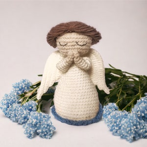 May include: A crocheted angel figurine with closed eyes and hands clasped in prayer. The angel is wearing a white dress with blue trim and has brown hair. The angel is surrounded by blue flowers.