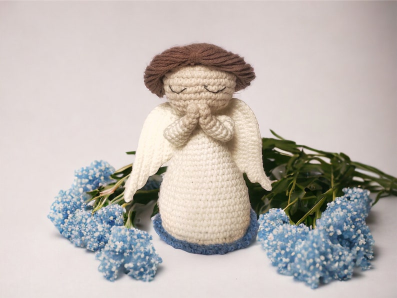 May include: A crocheted angel figurine with closed eyes and hands clasped in prayer. The angel is wearing a white dress with blue trim and has brown hair. The angel is surrounded by blue flowers.