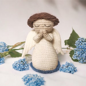 May include: A crocheted angel figurine with closed eyes, brown hair, and white wings. The angel is wearing a white dress with a blue base and is surrounded by blue flowers.