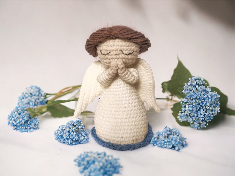 May include: A crocheted angel figurine with closed eyes, brown hair, and white wings. The angel is wearing a white dress with a blue base and is surrounded by blue flowers.